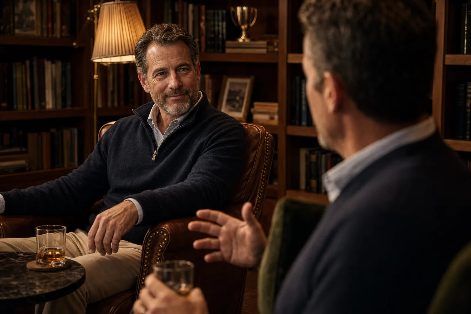 Two men in leather armchairs in conversation over whiskey in a book-lined library.